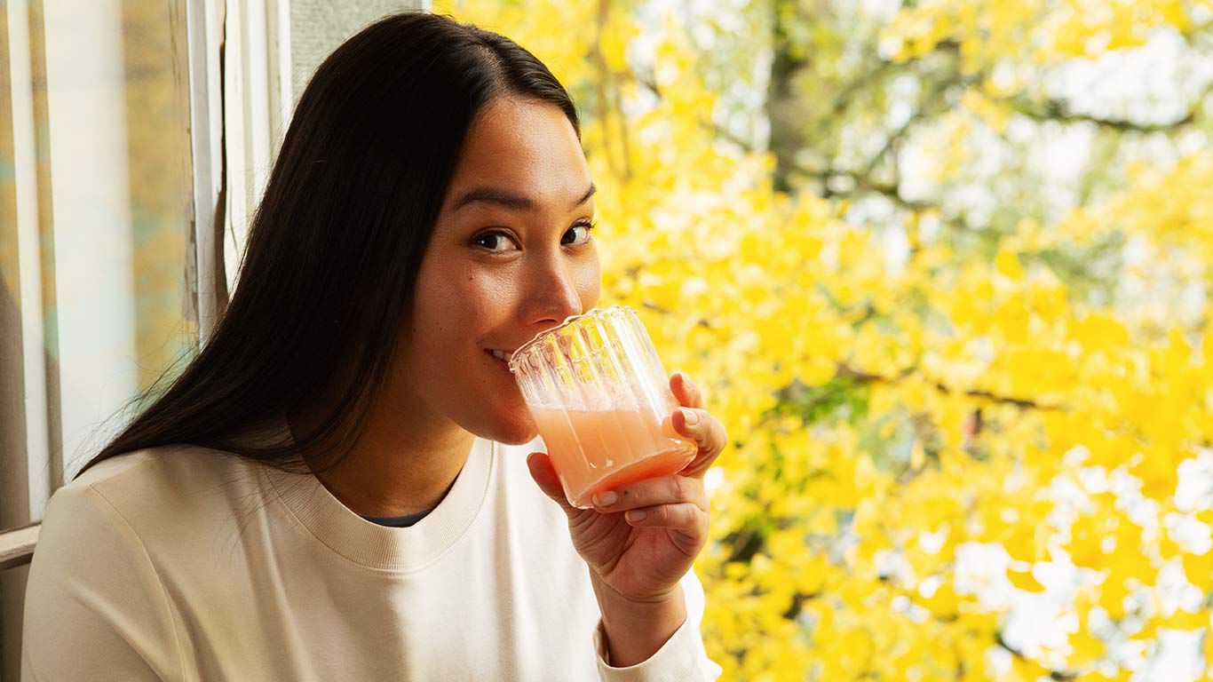 Vrouw zit voor het raam en drinkt uit een glas. 